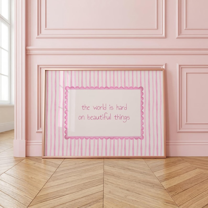 Peut inclure: Une &oelig;uvre d'art murale encadr&eacute;e rectangulaire avec un fond &agrave; rayures verticales rose clair et blanc. Au centre, un panneau rectangulaire blanc avec une bordure rose festonn&eacute;e affiche le texte : the world is hard on beautiful things.