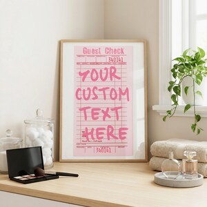 May include: A framed vertical wall art print featuring a pink restaurant guest check design with the text "GUEST CHECK" at the top and "YOUR CUSTOM TEXT HERE" in large pink letters, with the number "340141" printed at the top and bottom.