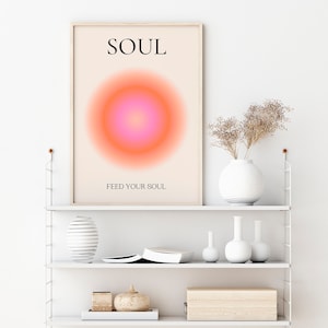 May include: A framed art print with the word "SOUL" at the top and "FEED YOUR SOUL" at the bottom. The artwork features a gradient circle in shades of pink and orange. The print is displayed on a white shelf with decorative vases and a wooden box.