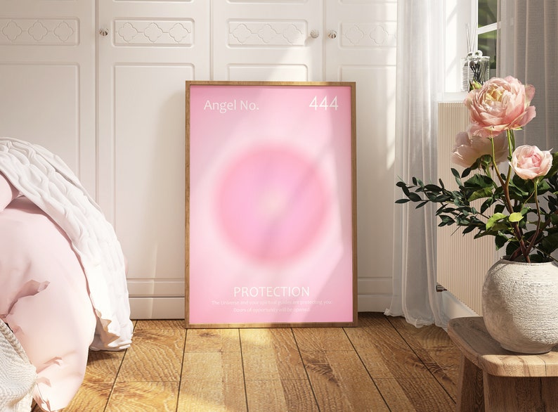 May include: A framed pink and white poster with the text "Angel No. 444 PROTECTION" and a pink circle in the center. The poster is hanging on a white wall in a room with a white bed and a vase of pink flowers.