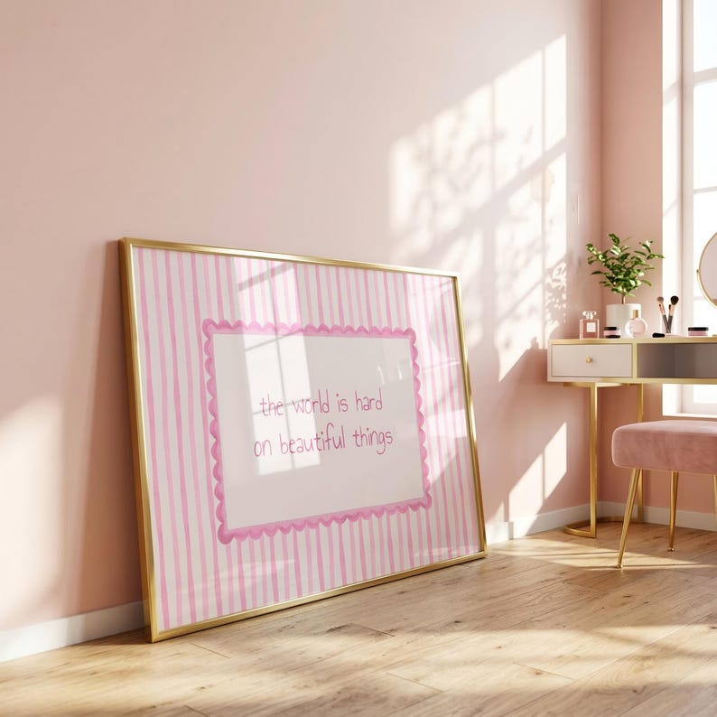 Peut inclure: Une &oelig;uvre d'art murale rectangulaire dans un cadre dor&eacute;, pr&eacute;sentant un panneau central blanc avec une bordure rose festonn&eacute;e et le texte "the world is hard on beautiful things" en rose, sur un fond &agrave; rayures verticales roses et blanches.