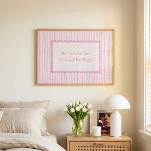 Peut inclure: Une impression murale rectangulaire dans un cadre en bois clair avec un fond &agrave; rayures verticales roses et blanches. Un rectangle blanc central avec une bordure rose festonn&eacute;e contient le texte : the world is hard on beautiful things.