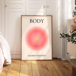 May include: A framed art print with the word "BODY" at the top and "NOURISH YOUR BODY" at the bottom. The artwork features a soft, blurred, circular design in shades of pink and yellow. The print is displayed in a room with a bed and flowers.