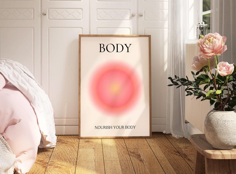 May include: A framed art print with the word "BODY" at the top and "NOURISH YOUR BODY" at the bottom. The artwork features a soft, blurred, circular design in shades of pink and yellow. The print is displayed in a room with a bed and flowers.