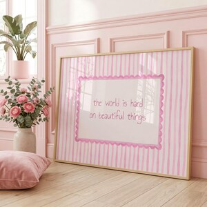 Peut inclure: Une impression encadr&eacute;e rectangulaire avec un panneau central blanc bord&eacute; d'une bordure rose festonn&eacute;e, sur un fond &agrave; rayures roses et blanches. Le texte "the world is hard on beautiful things" est imprim&eacute; en rose au centre du panneau.