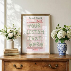 May include: A framed print of a pink guest check with red grid lines and text that reads Guest Check, 340141, and YOUR CUSTOM TEXT HERE in large green letters. The print is displayed in a gold frame on a wooden surface between two flower vases.