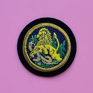 Könnte beinhalten: Ein gold gestickter Löwen-Patch mit einer Schlange und grünen Blättern auf einem schwarzen Filzgrund. Der Löwe ist von einer goldenen Litze umgeben.
