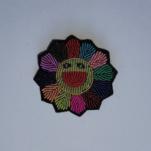 May include: A colorful embroidered flower patch with a smiling face in the center. The flower has petals in shades of pink, purple, blue, green, yellow, and black.