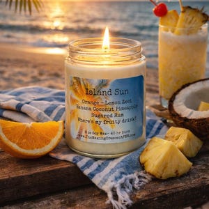 May include: A lit Island Sun scented candle in a glass jar with a label listing scents like orange, lemon zest, and pineapple. A cocktail with a cherry and pineapple garnish sits nearby, along with pineapple chunks and an orange slice.