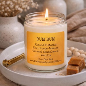 May include: A lit candle in a clear glass jar with a yellow label that reads "BUM BUM" and lists scents like Almond Pistachio and Vanilla. The candle sits on a white plate with a gold-colored handle, accompanied by caramel candies and decorative beads.
