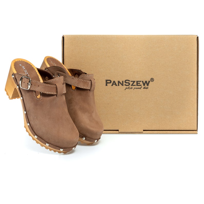 May include: A pair of brown leather clogs with a buckle strap and studded heels. The clogs are sitting on a brown cardboard box with the text "PANSZEW&reg;" and "jakiś podkład tekst" printed on it.