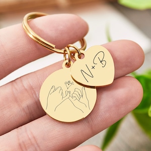 May include: Gold keychain with two charms. One charm is a heart with the text "N+B" and the other charm features two hands making a pinky promise with a small heart in the middle.