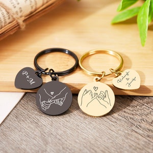 May include: Two matching keychains with a heart charm and a round charm with a line drawing of two hands holding pinkies. One keychain is black with the text "P+M" on the heart charm. The other keychain is gold with the text "Raleigh & Jennifer" on the heart charm.