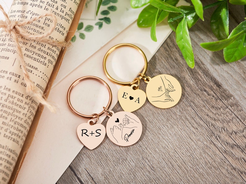 May include: Two gold keychains with heart and circle charms. The heart charms are engraved with the initials "E & A" and "R & S". The circle charms feature a line drawing of two hands intertwined with the word "Promise" on one charm.