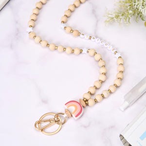 May include: A wooden bead lanyard with a gold-colored clasp and a rainbow charm. The lanyard features light-colored wooden beads and white beads with the name "Nicole" spelled out. The rainbow charm has pink, orange, and yellow colors.