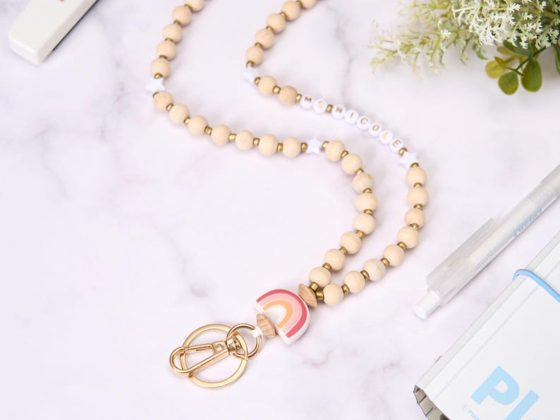 May include: A wooden bead lanyard with a gold-colored clasp and a rainbow charm. The lanyard features light-colored wooden beads and white beads with the name "Nicole" spelled out. The rainbow charm has pink, orange, and yellow colors.