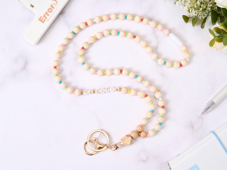 May include: A wooden bead lanyard with a gold-colored clasp and key ring. The lanyard features natural-colored wooden beads interspersed with colorful beads. The text "MSBAZIOW" is spelled out in white letter beads. A pen and notepad are in the background.
