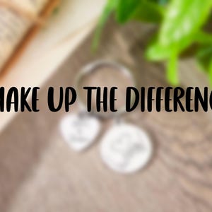 May include: A silver-toned metal keyring featuring a circular ring, a heart-shaped charm, and a circular charm. The text "MAKE UP THE DIFFERENCE" is prominently displayed in black lettering across the centre of the image over the charms.