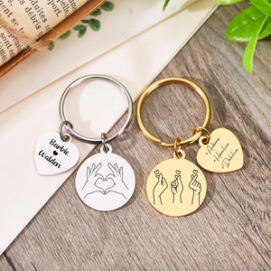 May include: Two silver keychains with heart-shaped charms. One keychain has the names "Barbie Walden" on the charm and the other keychain has the names "Ackers * Hemlia * Dahlia" on the charm. Both keychains have a round charm with a hand gesture design.