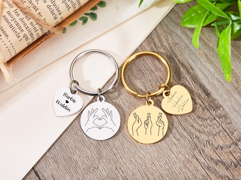 May include: Two silver keychains with heart-shaped charms. One keychain has the names "Barbie Walden" on the charm and the other keychain has the names "Ackers * Hemlia * Dahlia" on the charm. Both keychains have a round charm with a hand gesture design.