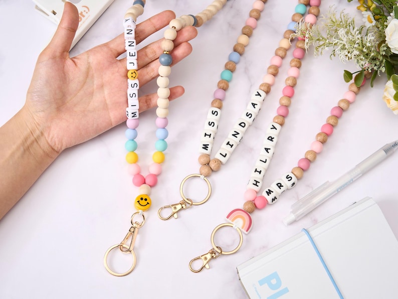 May include: Wooden bead lanyards with names and smiley faces. The lanyards feature a variety of pastel-colored beads, including pink, blue, yellow, and white. Each lanyard has a gold-colored clasp and a name spelled out in white letter beads.