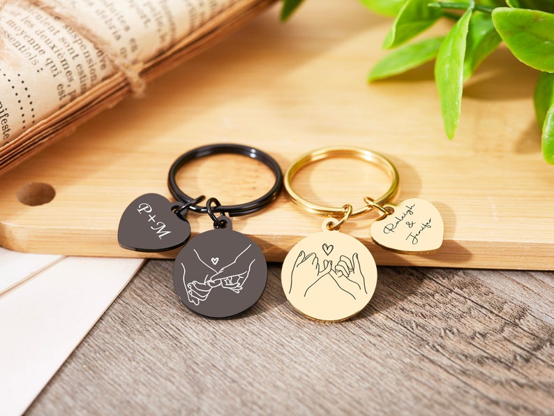 May include: Two matching keychains with a heart charm and a round charm with a line drawing of two hands holding pinkies. One keychain is black with the text "P+M" on the heart charm. The other keychain is gold with the text "Raleigh & Jennifer" on the heart charm.