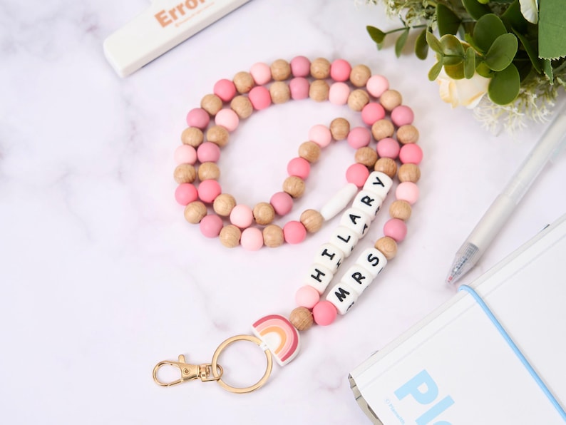 May include: A pink, tan, and white beaded lanyard with the name "HILARY MRS" spelled out in white letter beads. The lanyard has a gold-colored clasp and a rainbow charm. The word "Error" is visible on a white object in the background.