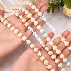 May include: Wooden bead necklaces with gold accents and colorful beads. Some necklaces feature white star-shaped beads and white letter beads spelling out names. The beads are strung together with a natural cord.