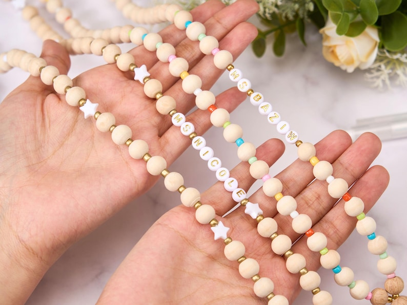 May include: Wooden bead necklaces with gold accents and colorful beads. Some necklaces feature white star-shaped beads and white letter beads spelling out names. The beads are strung together with a natural cord.