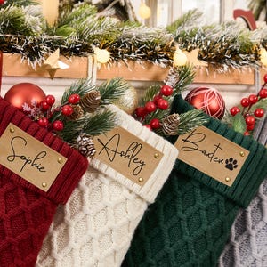 May include: Four personalised Christmas stockings with names "Sophie", "Ashley", "Baxter", and "Joslyn" on brown paper tags. The stockings are made of knitted fabric in red, white, green, and grey.