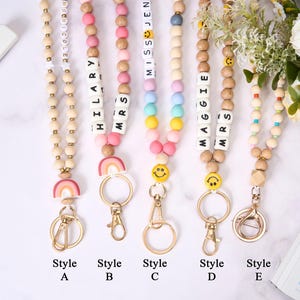 May include: A collection of beaded lanyards in various styles. Each lanyard features wooden beads, colorful accent beads, and a metal clasp. Some lanyards include names spelled out with letter beads, smiley faces, and rainbow charms. The styles are labeled A through E.