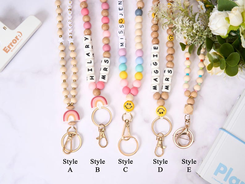 May include: A collection of beaded lanyards in various styles. Each lanyard features wooden beads, colorful accent beads, and a metal clasp. Some lanyards include names spelled out with letter beads, smiley faces, and rainbow charms. The styles are labeled A through E.