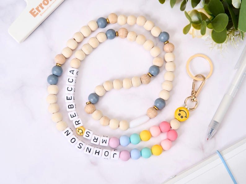 May include: A wooden bead lanyard with the name "REBECCA MRS JOHNSON" spelled out in white letter beads. The lanyard features light blue, natural wood, and rainbow-colored beads, and a gold-colored smiley face charm.