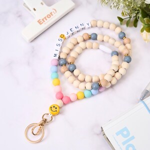May include: A wooden bead lanyard with a gold clasp and a smiley face charm. The lanyard features the name "Jenny" spelled out in white letter beads and a rainbow of pastel-colored beads. The word "Error:)" is printed on a white clip.