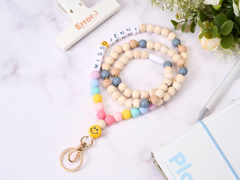 May include: A wooden bead lanyard with a gold clasp and a smiley face charm. The lanyard features the name "Jenny" spelled out in white letter beads and a rainbow of pastel-colored beads. The word "Error:)" is printed on a white clip.