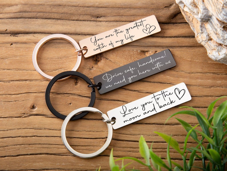 May include: Three silver keychains with engraved messages. The top keychain has a rose gold finish and reads "You are the greatest catch of my life". The middle keychain is black and reads "Drive safe, handsome! We need you here with us". The bottom keychain is silver and reads "Love you to the moon and back".