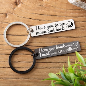 May include: Two silver keychains with black rings. One keychain has the text "I love you to the moon and back" with a crescent moon and stars. The other keychain has the text "I love you handsome I need you here with me" with a heart.