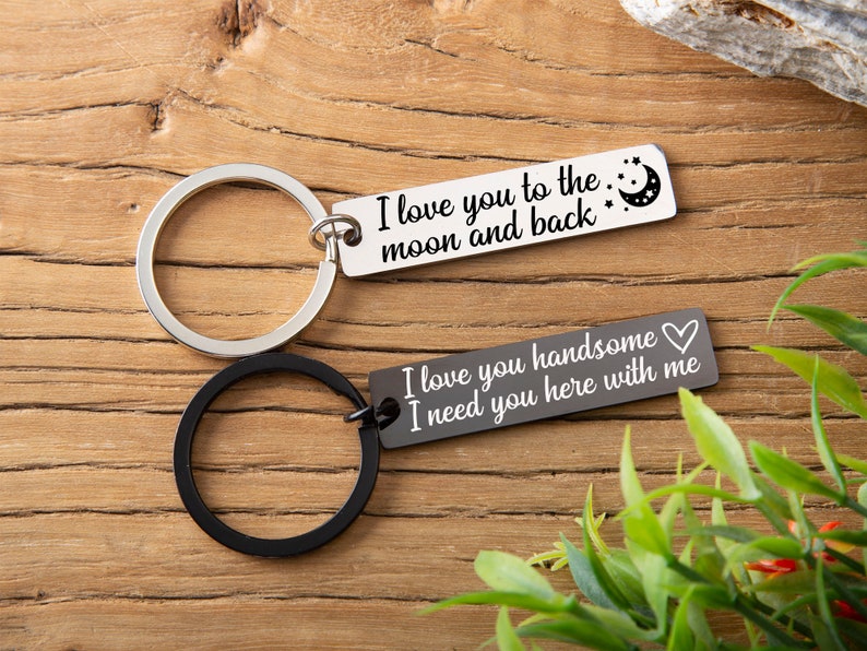 May include: Two silver keychains with black rings. One keychain has the text "I love you to the moon and back" with a crescent moon and stars. The other keychain has the text "I love you handsome I need you here with me" with a heart.