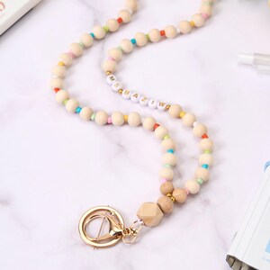 May include: A wooden bead lanyard with a gold clasp. The lanyard features natural wood beads interspersed with colorful beads. The word "Ms. Amazing" is spelled out in white and gold letter beads. A gold-colored swivel clasp and wooden geometric bead complete the design.