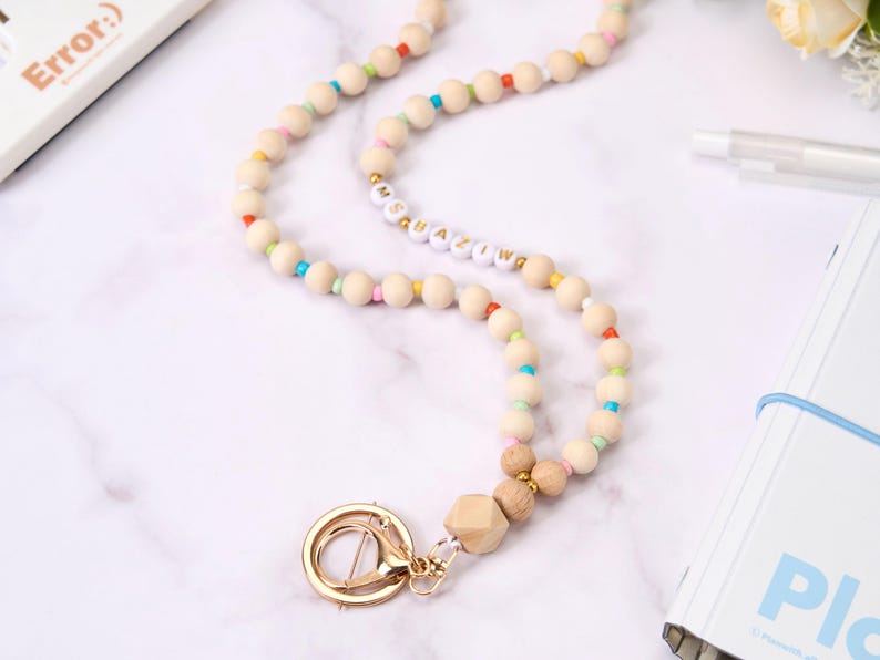 May include: A wooden bead lanyard with a gold clasp. The lanyard features natural wood beads interspersed with colorful beads. The word "Ms. Amazing" is spelled out in white and gold letter beads. A gold-colored swivel clasp and wooden geometric bead complete the design.