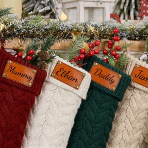 Peut inclure: Quatre chaussettes de Noël en tricot avec des étiquettes de nom en cuir personnalisées. Les chaussettes sont rouge, blanche, verte et crème. Les noms sur les étiquettes sont "Mummy", "Ethan", "Daddy" et "James".