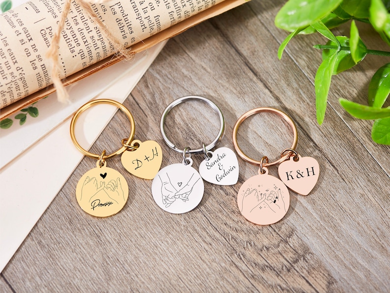 May include: Three gold, silver, and rose gold keychains with heart-shaped charms and round charms featuring a pinky promise design. The charms are engraved with the names "D+H", "Sandra & Godwin", and "K&H".