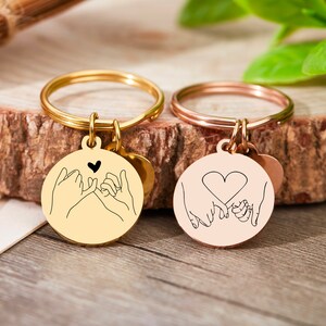 May include: Two gold keychains with a small round charm featuring a line drawing of two hands intertwined with a heart. One keychain is gold and the other is rose gold.