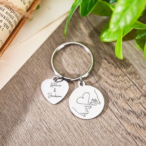 May include: A silver keychain with two charms. One charm is a heart-shaped tag with the names "Kathie & Jackson" engraved on it. The other charm is a round tag with a line drawing of two hands holding a heart.