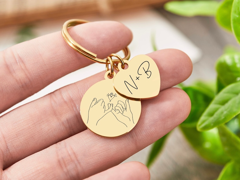 May include: Gold keychain with two charms. One charm is a heart with the text "N+B" and the other charm features two hands making a pinky promise with a small heart in the middle.