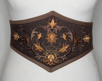 May include: A brown corset belt with intricate gold floral embroidery. The belt has a wide, contoured shape and is displayed on a mannequin. The embroidery features detailed leaves and swirling patterns, creating a decorative accessory.
