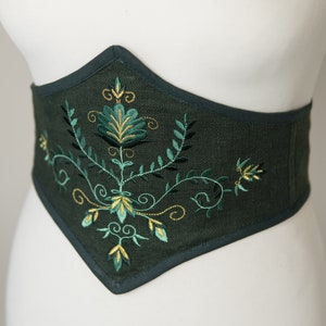 May include: A dark green linen corset with a floral embroidery design in shades of green and gold.