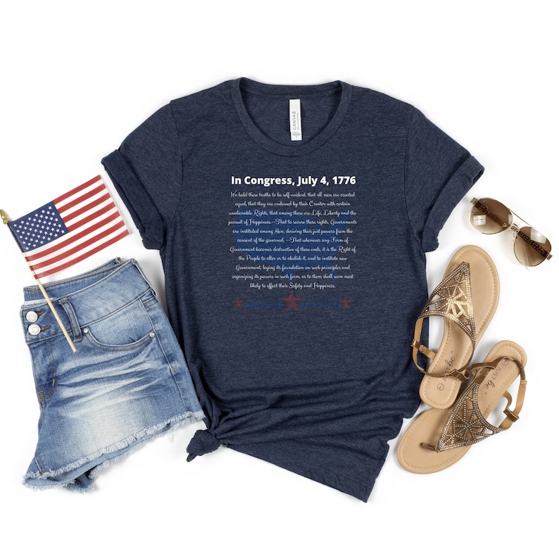 May include: A navy blue t-shirt featuring the text "In Congress, July 4, 1776" and the Declaration of Independence in white. The shirt is styled with denim shorts, a small American flag, and gold sandals.
