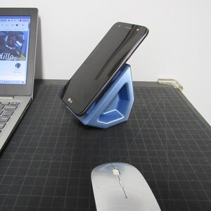 May include: A blue 3D printed phone stand holds a black smartphone with the LG logo visible. A silver wireless mouse and a silver laptop are also on the black surface.
