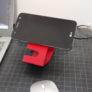 Puede incluir: Un soporte de teléfono de plástico rojo con un teléfono inteligente negro descansando en la parte superior. El teléfono está conectado a un cable USB.